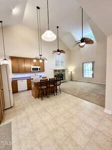 Open kitchen and living area with tiled and carpeted floors, brown cabinets, an island with three chairs, a fireplace, ceiling fans, pendant lights, and a window letting in natural light.