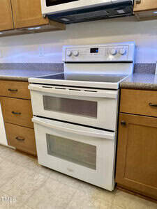 A white double oven stove with a digital display is installed between wooden cabinets and beneath a mounted microwave in a kitchen with beige countertops and tile flooring.