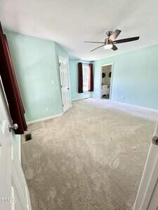 A bedroom with light blue walls, beige carpet, dark red curtains, a ceiling fan with a light, and an open door leading to a bathroom. The room is empty and well-lit by natural light.