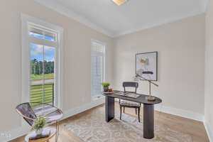 A home office with a modern black desk, chair, desk lamp, and decorative items. There are two tall windows, light-colored walls, wood flooring, a geometric rug, and abstract wall art. A clear chair sits in the corner.