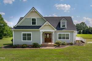 A light grey two-story house at 274 S Chaucer Way, Kittrell, with white trim, a dark shingle roof, and a front porch with wooden double doors. The home sits among a lawn, shrubs, and trees under a partly cloudy sky.