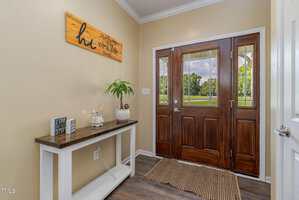 A small foyer with beige walls, a dark wooden front door with glass panels, a white console table holding decor items and a potted plant, and a brown woven rug on a wood floor. A wooden wall sign hangs above the table.