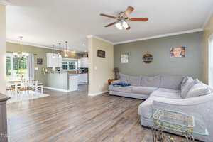 A bright living room with wood floors, a large gray sectional sofa, ceiling fan, and adjacent dining area. The kitchen with white cabinets and pendant lights is visible in the background. Family photos hang on the walls.