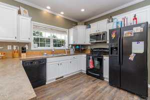 A kitchen with white cabinets, black appliances, a brown countertop, a double sink under a window, and wooden flooring. Small kitchen items, magnets, and papers are visible on the counters and refrigerator.