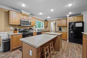 A kitchen with wood cabinets, black appliances, beige countertops, a center island with two stools, and recessed ceiling lights. The room has tan walls, white trim, and a window above the sink.