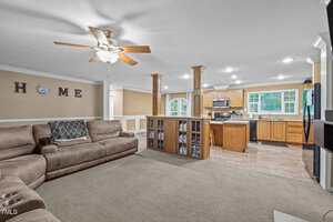 An open living room with a beige sectional sofa, ceiling fan, and carpeted floor leads into a kitchen with wood cabinets, stainless steel appliances, and a central island with built-in shelving.
