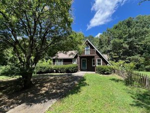 Nestled at 162 Black Horse Lane, Kittrell, this dark brown A-frame house features a small balcony and green front door, surrounded by trees and grass. A paved driveway leads to the entrance, with a wooden fence lining the right side.