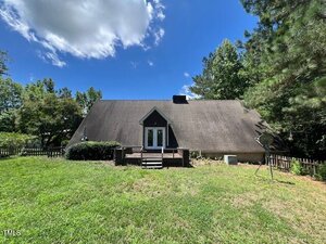 A-frame house with a steep roof, central double doors, small porch with steps, surrounded by green grass, trees, and a wooden fence, under a blue sky with scattered clouds.