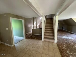 A room with tiled flooring and beige walls leads to a carpeted staircase going up. The adjoining rooms have unfinished or damaged flooring, and natural light enters from windows on the left.