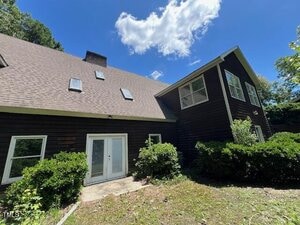 A two-story dark brown house with large windows, skylights, and a steep roof is shown under a blue sky with scattered clouds. There are bushes and a small concrete patio by the double doors.