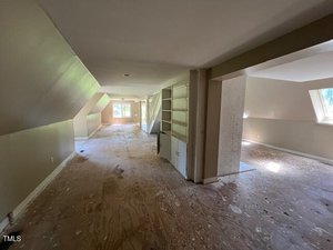 A long, empty room with unfinished plywood floors, beige walls, built-in shelves, and several windows letting in natural light. The space appears to be under renovation or in need of flooring.