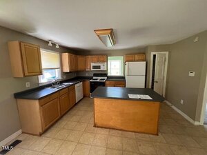 A kitchen with light brown cabinets, black countertops, a white refrigerator, stove, microwave, and dishwasher. There is an island in the center, beige tile floor, and two windows letting in natural light.