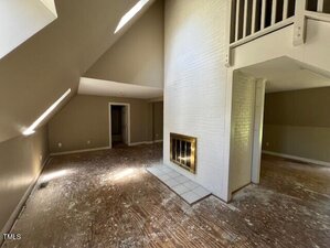 Unfurnished room with slanted ceilings, a white brick fireplace, bare floors with scattered debris, and doorways leading to other areas. Upper level railing is visible above the fireplace. Natural light comes from above.