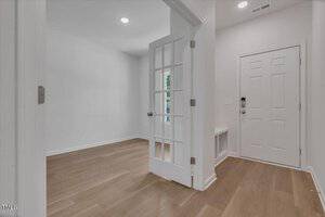 A bright entryway with light wood floors, white walls, a white front door, and a glass-paned door leading to an adjacent room. Recessed ceiling lights and a built-in bench with cubbies are visible.