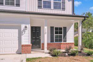 A modern two-story house with white siding, black shutters, a black front door, a covered porch with white columns, and a garage on the left. Some small shrubs and a grass lawn are in front of the porch.