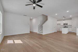 Open-plan living area with light wood flooring, white walls, a ceiling fan, and recessed lighting. Kitchen with white cabinets, oven, and microwave is visible in the background. Stairs lead to the upper floor.
