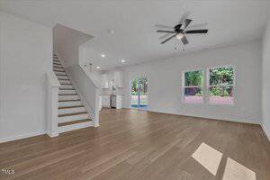 A spacious, empty living room with light wood flooring, white walls, a ceiling fan with lights, stairs to the left, and large windows and a glass door overlooking a backyard with trees. The kitchen is visible in the background.