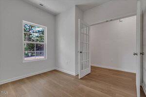 A small empty room with light wood flooring, white walls, a single window with a view of trees, and a French door leading to another room. The space is well-lit with natural light.