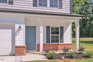 The front entrance of a house with white siding, brick accents, a blue door, two windows with black shutters, house number 114, a porch light, and a small landscaped area with shrubs.