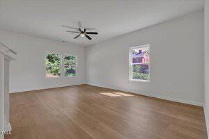 Empty, unfurnished room with light wood flooring, white walls, two large windows showing greenery and a neighboring house, and a ceiling fan with a light.
