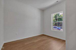 A small, empty room with white walls, light wood flooring, a single window with a view of green trees and a parked blue vehicle outside during daytime.
