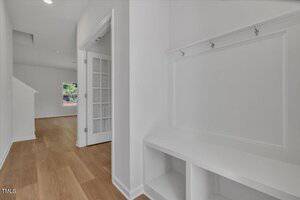A hallway with light wood flooring, white walls, a built-in white bench with storage and hooks, a set of French doors, and a view into another room with a window.