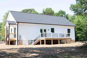 Single-story white house with black roof, small deck and stairs leading to a backyard area. There are several windows and a glass door opening onto the deck, surrounded by trees and natural landscaping.