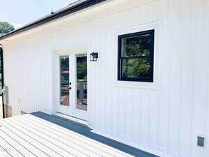 A white exterior wall with vertical siding features a black-framed window and glass double doors. A black outdoor wall light is mounted beside the doors. The deck below is light gray. Trees are visible in the reflection.