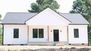 A white single-story house with a gable roof, black-framed windows, wooden porch columns, and a black front door, set in front of tall green trees.