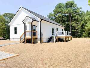 A white, single-story house with black trim, a small covered front porch, and stairs leading to a wooden deck. The house sits on a dirt lot with sparse vegetation and is surrounded by trees.
