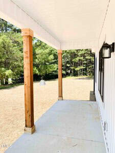 A covered porch with a concrete floor, wooden support columns, and a white ceiling. The porch overlooks a grassy yard with trees in the background. The exterior wall of the house is white with black-trimmed windows.