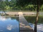 A wooden dock at 9961 Lakeside Terrace extends over a calm lake, leading to a larger platform at the end. Surrounded by trees with green foliage in Bullock, the water beautifully reflects the lush scenery.
