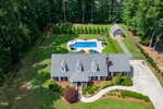 Aerial view of a brick house with a gray roof at 4135 Somerset Drive, Oxford, featuring a landscaped yard, curved driveway, fenced backyard, in-ground pool with patio area, detached garage, all nestled among trees.