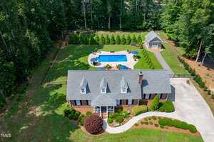 Aerial view of a brick house with a gray roof at 4135 Somerset Drive, Oxford, featuring a landscaped yard, curved driveway, fenced backyard, in-ground pool with patio area, detached garage, all nestled among trees.
