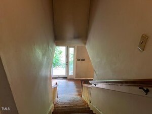 View from the top of a carpeted staircase with wooden handrails, leading down to a landing with a glass door that opens to an outdoor area with greenery visible through the door. Walls appear plain and slightly worn.