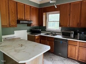 A kitchen with wooden cabinets, a dirty white countertop island, stainless steel dishwasher, microwave, toaster oven, and a window above the sink. The walls are green and the floor is tiled.
