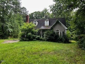 A gray house with two dormer windows and a brick chimney sits surrounded by tall trees and overgrown greenery. The roof has visible wear, and the yard is covered with unruly grass and bushes.