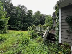 An overgrown yard with tall grass and dense vegetation surrounds a weathered wooden deck attached to a house. The deck's railing is broken, and vines are growing up the side of the house. Trees are visible in the background.
