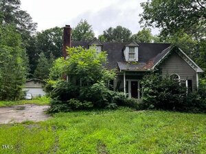 A two-story, gray house with a steep roof and chimney is partially obscured by overgrown bushes and trees. The house sits on a grassy lot with a driveway leading to a detached garage in the background.