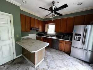 A kitchen with green walls, wooden cabinets, a marble-topped island, stainless steel appliances, and a ceiling fan. The floor is tiled, and there is a window above the sink.