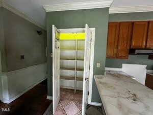 A pantry with double doors open, revealing empty white shelves. The pantry interior has a yellow-tinted light. Adjacent are green walls, wooden cabinets, and a stained countertop in a kitchen area.