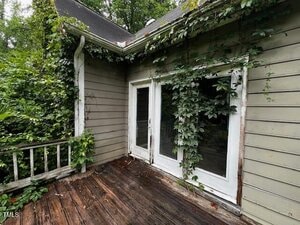 A weathered wooden deck with overgrown vines climbing on the siding and around the white-framed glass doors of a house; the siding is worn and paint is peeling in places. Trees and dense greenery are visible in the background.