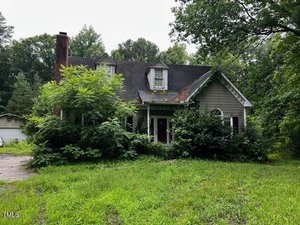 Nestled at 3573 River View Circle in Oxford, this two-story house with a dark roof and dormer windows is partially hidden by overgrown trees and bushes. The unkempt yard and weathered exterior show it needs some maintenance.