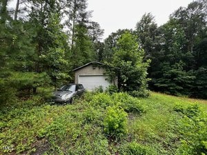 A small white garage surrounded by dense green trees and overgrown bushes, with a dark car parked in front on a patch of grass. The sky is overcast.