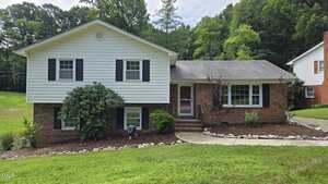 This two-story split-level house at 505 Forest Road, Oxford, features white siding above red brick, a front yard with green grass, shrubs, and a curved walkway leading to the door. Tall trees stand in the background.