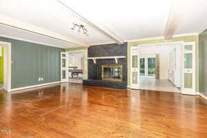 A spacious living room with hardwood floors, green paneled walls, a black brick fireplace with a mirrored insert, and glass double doors leading to a bright sunroom with large windows.