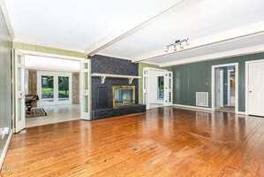 A spacious living room with dark green walls, wood flooring, a black brick fireplace with a brass-trimmed glass door, large windows, and open doorways leading to adjacent rooms and a view of a backyard.