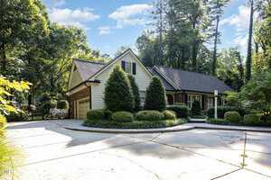 A single-story house with beige siding and brick accents sits among tall trees. The front yard features manicured shrubs and a circular driveway leading to a two-car garage. The sky is partly cloudy.
