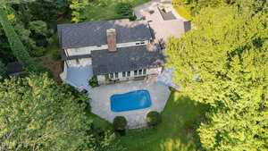 A two-story house with a shingled roof is shown from above, featuring a backyard with a small in-ground swimming pool, a patio area, and surrounded by green trees and grass.