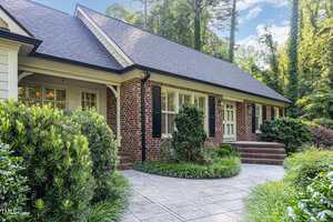 A single-story brick house with a dark shingle roof, black shutters, white-framed windows, and a covered front porch. There are steps leading to the entrance and a landscaped walkway surrounded by shrubs and greenery.
